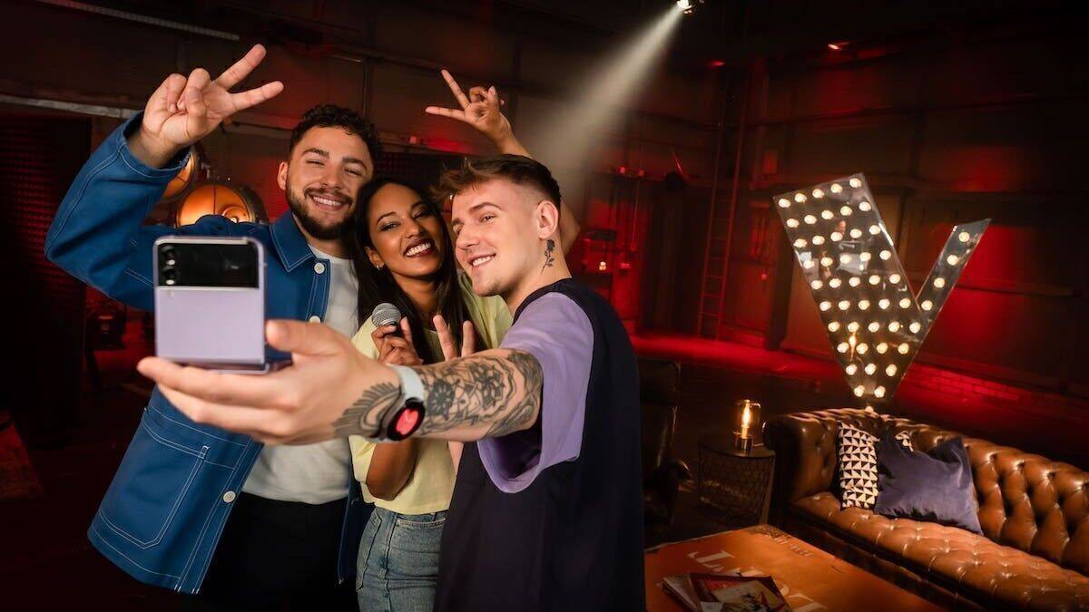 Zwei Männer und eine Frau machen ein Selfie mit einem Handy von Samsung, im Hintergrund ist das Logo von "The Voice of Germany" zu sehen.