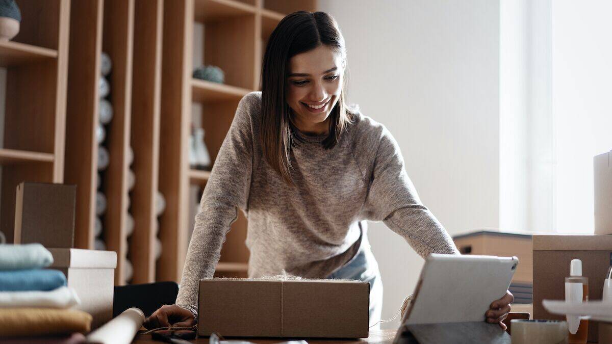 Eine Frau packt ein Paket, gleichzeitig schaut sie auf ein Tablet.